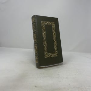 May include: A dark green hardcover book with gold detailing. The front cover features a gold rectangular frame with ornate designs. The spine has gold lettering. The book is displayed on a clear stand.