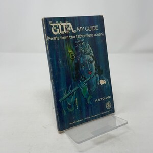Puede incluir: Libro antiguo titulado "GITA MY GUIDE" con una cubierta azul que presenta una ilustración de una figura tocando una flauta. El texto incluye "Pearls from the fathomless ocean" y el nombre del autor, P.S. Tolani. El libro se exhibe en un soporte acrílico transparente.