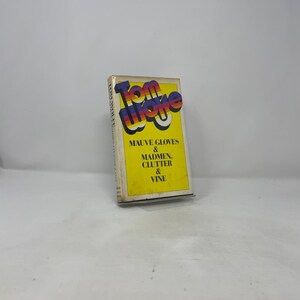 Könnte beinhalten: Ein Vintage-Taschenbuch mit dem Titel "Tom Wolfe" und dem Text "Mauve Gloves & Madmen, Clutter & Vine" auf gelbem Hintergrund. Der Titel ist in einer farbenfrohen Retro-Schrift.