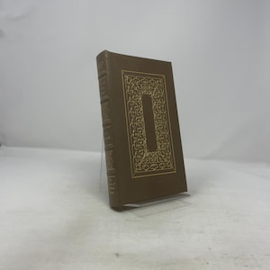 Op de afbeelding: Een hardcover boek met een bruine kaft en gouden decoratieve accenten. De kaft heeft een grote gouden letter "I" en sierlijke patronen. Het boek wordt getoond op een heldere acryl standaard, wat duidt op een verzamelobject of decoratief item.