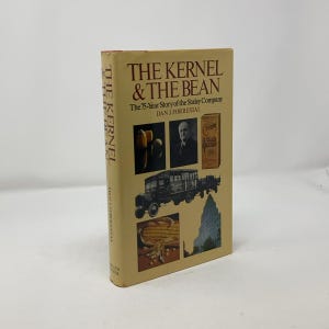 以下が含まれることがあります： ダニ・フォレストール著「ザ・カーネル＆ザ・ビーン：ザ・ステーリー・カンパニー75年の物語」というタイトルの黄色のハードカバーの本。表紙には、トウモロコシ、スーツを着た男性、トラック、建物の画像が掲載されています。