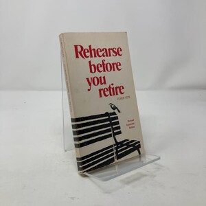 May include: A vintage book titled "Rehearse before you retire" by Elmer Otte. The book cover is off-white with red text and a black and white illustration of a park bench and a bird. The book is on a clear display stand.