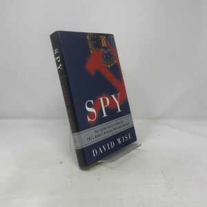 May include: A hardcover book titled "SPY" by David Wise. The cover is navy blue with the title in large white letters and a red arrow graphic. The subtitle reads "The Inside Story of how the FBI's Robert Hanssen Betrayed America."