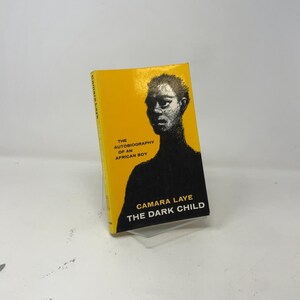 May include: A hardcover book titled "The Dark Child" by Camara Laye. The book has a bright yellow cover with black text and a black silhouette of a person's face. The book is displayed on a clear stand.