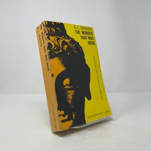 May include: A hardcover book titled "The Wonder That Was India" by A.L. Basham. The book has a yellow cover with a black illustration of a face. The spine of the book is orange.