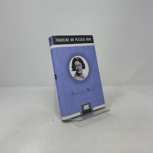 May include: A hardcover book titled "Simone Weil" by Francine Du Plessix Gray. The book has a light purple cover with a portrait of a woman in a white circle. The book is propped up on a clear plastic stand.