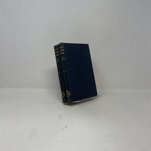 May include: Two hardback books with dark blue covers are displayed on a clear stand. The spines of the books are visible, with gold lettering and decorative elements. The books are titled "Jonas, A. & B." and "Jonas, A. & B. Vol. 1 & 2".