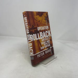 Peut inclure: Un livre à couverture rigide intitulé "Operation Rollback" de Peter Grose. La couverture est marron et orange avec du texte blanc. Le titre et le nom de l'auteur sont bien visibles. Le livre est présenté sur un présentoir transparent.