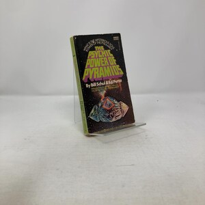 May include: A vintage paperback book titled "The Psychic Power of Pyramids" by Bill Schul & Ed Pettit. The cover features a black background with green and yellow text and an illustration of a geometric shape. The book is displayed on a clear stand.