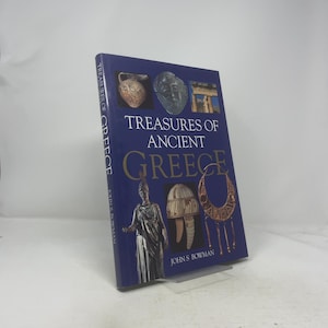 Puede incluir: Un libro de tapa dura titulado "Treasures of Ancient Greece" con una cubierta azul oscuro. La cubierta presenta imágenes de artefactos griegos y el nombre del autor, John S. Bowman. El libro se muestra en un soporte transparente.