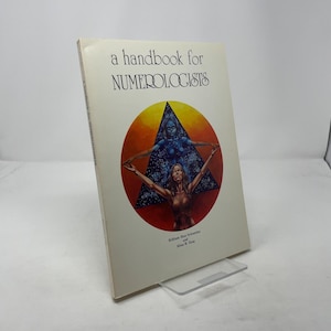 May include: A hardcover book titled "a handbook for NUMEROLOGISTS" with a colorful illustration. The cover art features two figures within a circle, one blue and one nude, with arms outstretched. The book is propped up on a clear plastic stand.