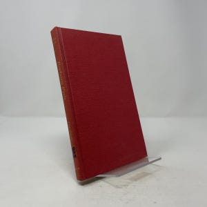 May include: A hardcover book with a red cover, titled "Barbara Frum" in gold lettering. The book is propped up on a clear acrylic stand, showcasing its spine and cover. The book's cover has a textured appearance.