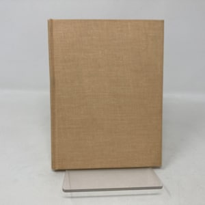 May include: A hardcover book with a tan linen cover. The book is standing upright on a clear acrylic stand. The cover has a subtle texture and a simple, classic design. The book's spine is visible, indicating a bound format.