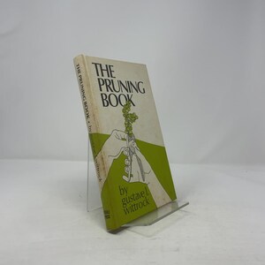 May include: A vintage book titled "The Pruning Book" by Gustave L. Wittrock. The cover features a green and white design with an illustration of pruning shears cutting a branch of grapes. The book is displayed on a clear stand.
