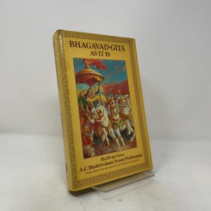Peut inclure: Un livre à couverture rigide jaune intitulé Bhagavad-Gita As It Is par A.C. Bhaktivedanta Swami Prabhupada. La couverture présente une illustration centrale d'un char tiré par des chevaux blancs avec un dais rouge, encadré par une bordure dorée.