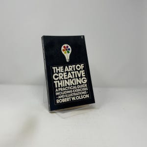May include: A hardcover book titled "THE ART OF CREATIVE THINKING" with a black cover. The cover features a white lightbulb graphic with colorful segments and white text. The book is a practical guide including exercises and illustrations by Robert W. Olson.