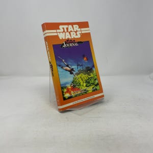 Puede incluir: Un libro naranja de Star Wars Journal con rayas y texto blancos. La portada presenta una escena de batalla espacial con cazas X-wing y un cielo púrpura. El libro se muestra en un soporte transparente.