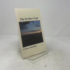 May include: A softcover book titled The Ox-Bow Trail: A Texas Adventure by Wayne Leavitt. The cover features a landscape photograph of a sunset over hills and trees, with black text printed on a cream-coloured background, displayed on a stand.