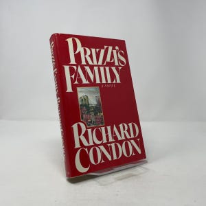 Puede incluir: Un libro de tapa dura rojo titulado "Prizzi's Family" de Richard Condon. El libro tiene letras blancas y una pequeña imagen de un edificio. El libro está de pie.