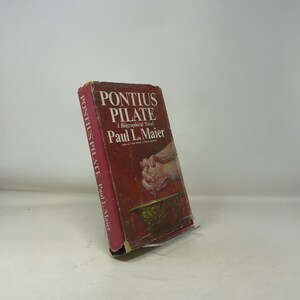 May include: A red hardcover book titled "Pontius Pilate" by Paul L. Maier. The cover features white text and an image of clasped hands. The book is displayed on a clear stand.
