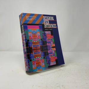 May include: A hardcover book titled Designing User Interfaces by James E. Powell. The cover features a dark blue background with a central graphic of colorful, pixelated geometric shapes and orange diagonal stripes at the top left corner.
