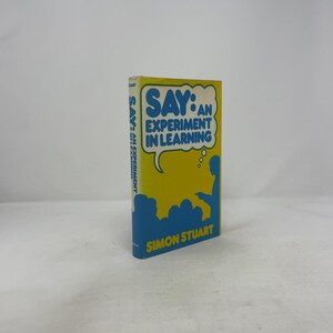 May include: A book titled "SAY: An Experiment in Learning" by Simon Stuart. The cover is yellow with blue and white text and graphics. The book's spine also features the title. The book is standing upright.