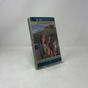 May include: A hardcover book titled "Ivanhoe" by Sir Walter Scott. The cover features a scene with a woman in a crown and a man on a horse, with a castle in the background. The book is propped up on a clear stand.