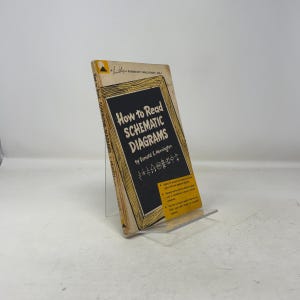 May include: A vintage book titled "How to Read Schematic Diagrams" by Donald E. Herrington. The book has a black and yellow cover with the title in large white letters. The book is propped up on a clear plastic stand.