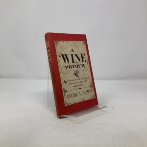 May include: A red hardcover book titled "A Wine Primer" by André L. Simon, displayed on a clear acrylic stand. The book's cover features ornate text and a decorative border. The book is a guide for beginners on how to buy, keep, and serve wine.