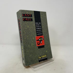 May include: A hardcover book titled Last Call by Tim Powers with a dark green speckled cover. The front features a black vertical stripe, a red square with a black Eye of Horus symbol, and text describing the book as a tale of gambling and fate.