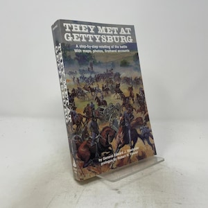 Könnte beinhalten: Ein gebundenes Buch mit dem Titel They Met at Gettysburg, das ein detailliertes historisches Gemälde einer Schlacht des Bürgerkriegs mit Soldaten zu Pferd und Kanonen zeigt. Das Cover enthält den Untertitel A step-by-step retelling of the battle.