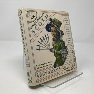 May include: A book titled "An Uncommon Scold" with an illustration of a woman in a green dress and a hat holding a fan of daggers. The book is by Abby Adams and features the text "1,000 Suitable Quotes on Subjects from the Period."