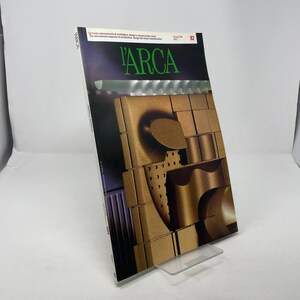 May include: A magazine titled "L'ARCA" with a green logo and the number 82. The cover features a gold-colored, abstract architectural design. The magazine is displayed on a clear plastic stand.