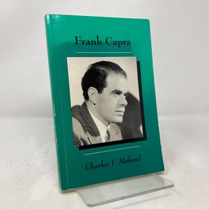 Puede incluir: Un libro de tapa dura verde con el título "Frank Capra" y el nombre del autor "Charles J. Maland" impreso en la portada. Una foto en blanco y negro de Frank Capra aparece en la portada.