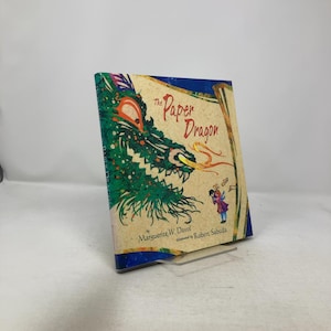 May include: A hardcover book titled "The Paper Dragon" by Marguerite W. Davol, illustrated by Robert Sabuda. The cover features a green and orange dragon illustration. A small fairy figure is also visible on the cover.