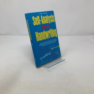 Può includere: Un libro blu intitolato "Self-Analysis for Your Handwriting" di Albert E. Hughes. Il titolo del libro è in giallo, con testo aggiuntivo in bianco e rosso. Il libro è esposto su un supporto di plastica trasparente.