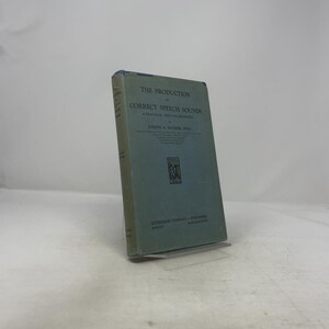 May include: A vintage book titled "The Production of Correct Speech Sounds" by Joseph A. Molder, Ph.D. The book has a light blue cover with dark blue text and a small illustration. The book is standing upright on a clear stand.