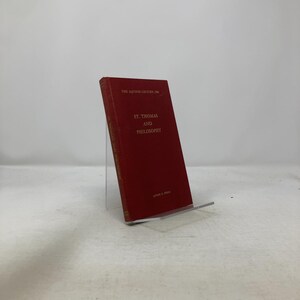 May include: A red hardcover book titled "St. Thomas and Philosophy" with gold lettering. The book is propped up on a clear acrylic stand. The title is centered on the cover, with additional text at the top and bottom.