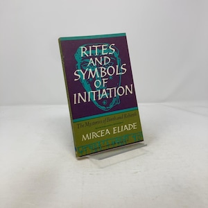 May include: A hardcover book titled "Rites and Symbols of Initiation" by Mircea Eliade. The book has a purple cover with teal and white text and a teal illustration of a face. The book is displayed on a clear stand.