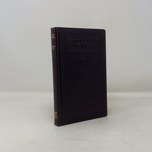 May include: A hardcover book titled "Modern Business" with gold lettering on a dark brown cover. The book has a textured surface and is standing upright. The spine also has gold lettering.