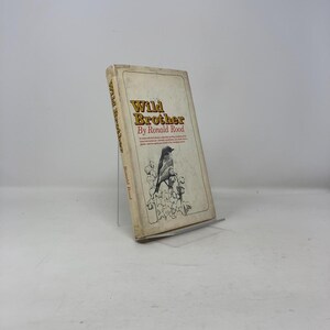 Puede incluir: Un libro antiguo titulado "Wild Brother" de Ronald Rood. La portada presenta una ilustración de un pájaro y texto en dorado y rojo sobre un fondo crema. El libro está apoyado en un soporte transparente sobre un fondo blanco.