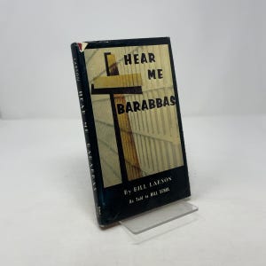 May include: A hardcover book titled "Hear Me Barabbas" by Bill Larson. The book cover features a graphic design with the title in bold letters and a cross-like image. The book is displayed on a clear acrylic stand.