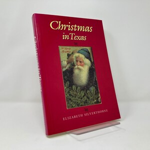 Pode incluir: Um livro de capa dura vermelho intitulado "Christmas in Texas" de Elizabeth Silverthorne. A capa apresenta uma ilustração vintage do Papai Noel com uma bolsa vermelha e uma árvore verde.