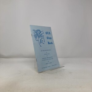 May include: A light blue "CEA Chap Book" with a unicorn illustration. The book's cover features text about mythology and a bibliography of myth in literature. The book is propped up on a clear stand.