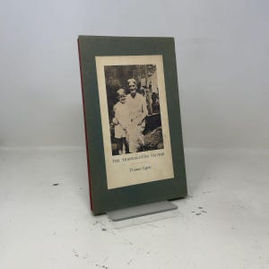 May include: A hardback book titled The Thanksgiving Visitor by Truman Capote. The cover is a muted dark green with a central cream-coloured rectangular inset featuring a black and white photograph of a child and an adult standing outdoors.