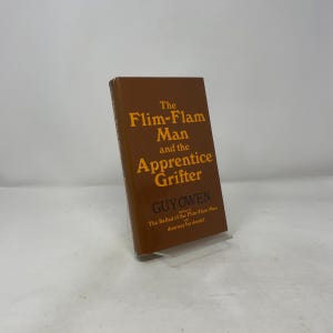 May include: A hardcover book titled "The Flim-Flam Man and the Apprentice Grifter" by Guy Owen. The book has a brown cover with gold lettering and is displayed on a clear stand. The book's title and author are clearly visible.