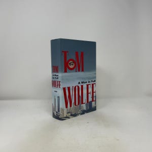以下が含まれることがあります： トム・ウルフ著『A Man in Full』のハードカバー本。ダストジャケットには青い空の背景、下部に都市の風景、大きな赤い文字が描かれています。TomのOの文字には小さな目の画像が含まれています。出版社FSGのロゴが印刷されています。