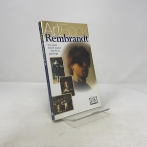 May include: A hardcover art book titled "ArtBook Rembrandt" featuring a portrait of Rembrandt. The book has a gold cover with the title in large letters and includes images of his paintings. The book is displayed on a clear stand.