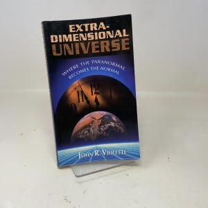 May include: A hardcover book titled "Extra-Dimensional Universe" by John R. Violette. The cover features a blue and black gradient with a clock face and a planet. The text "Where the paranormal becomes the normal" is also on the cover.