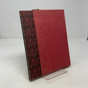 May include: Two antique books. One book has a red cover, and the other has a patterned cover with red and black floral designs. The books are propped up on a clear plastic stand against a white background.